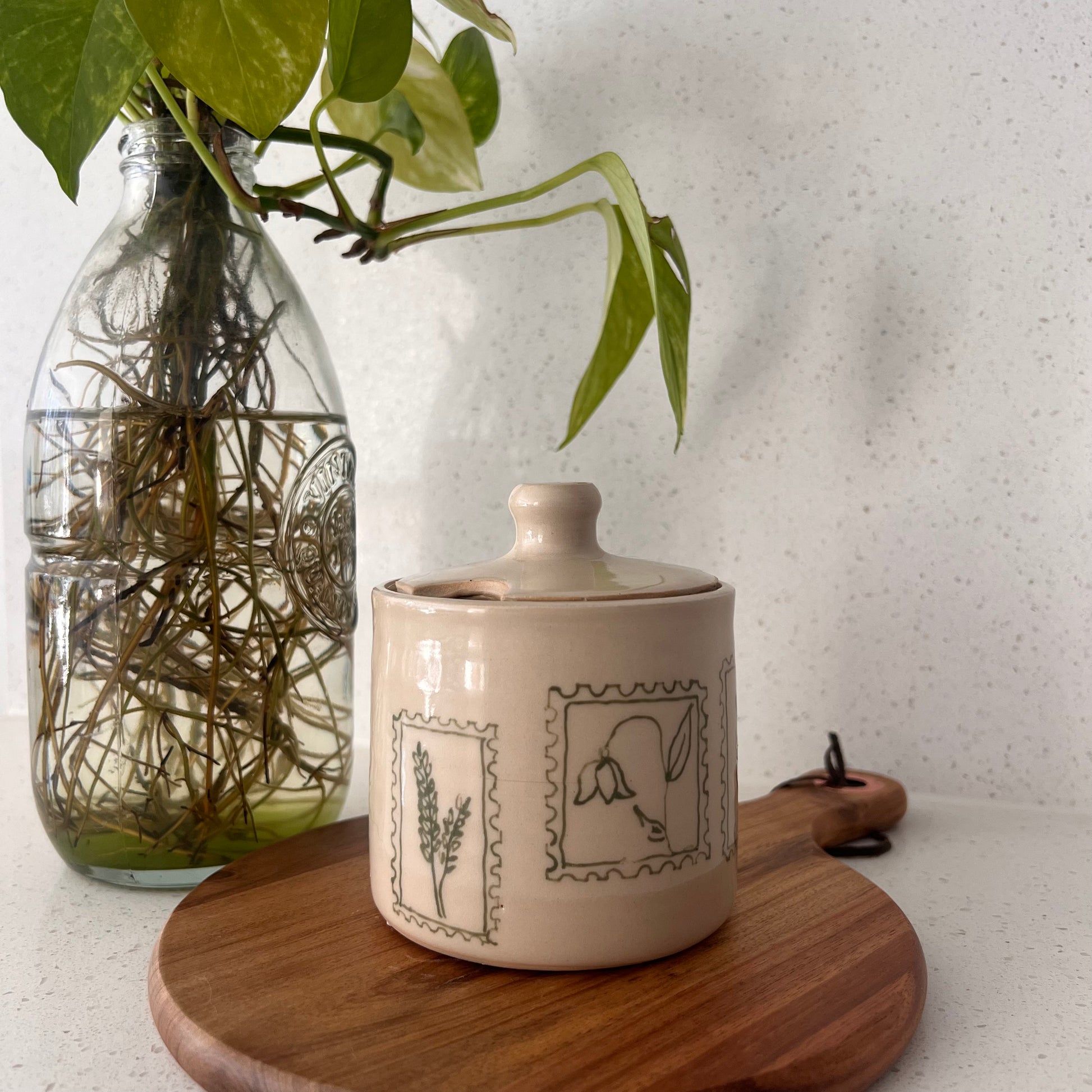 Ceramic jar with decorative elements on a wooden surface next to a glass vase with greenery.