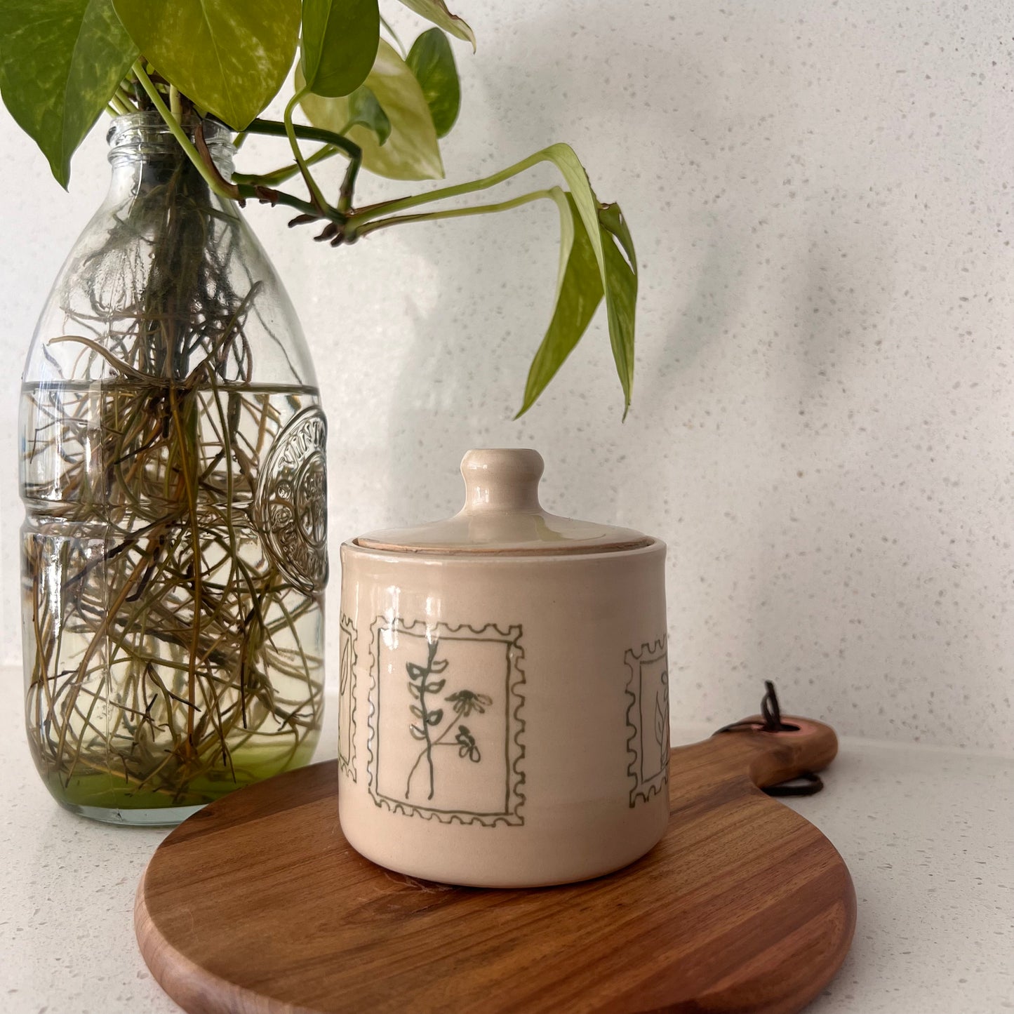 Ceramic jar with decorative design on a wooden board with a glass vase containing greenery in the background.