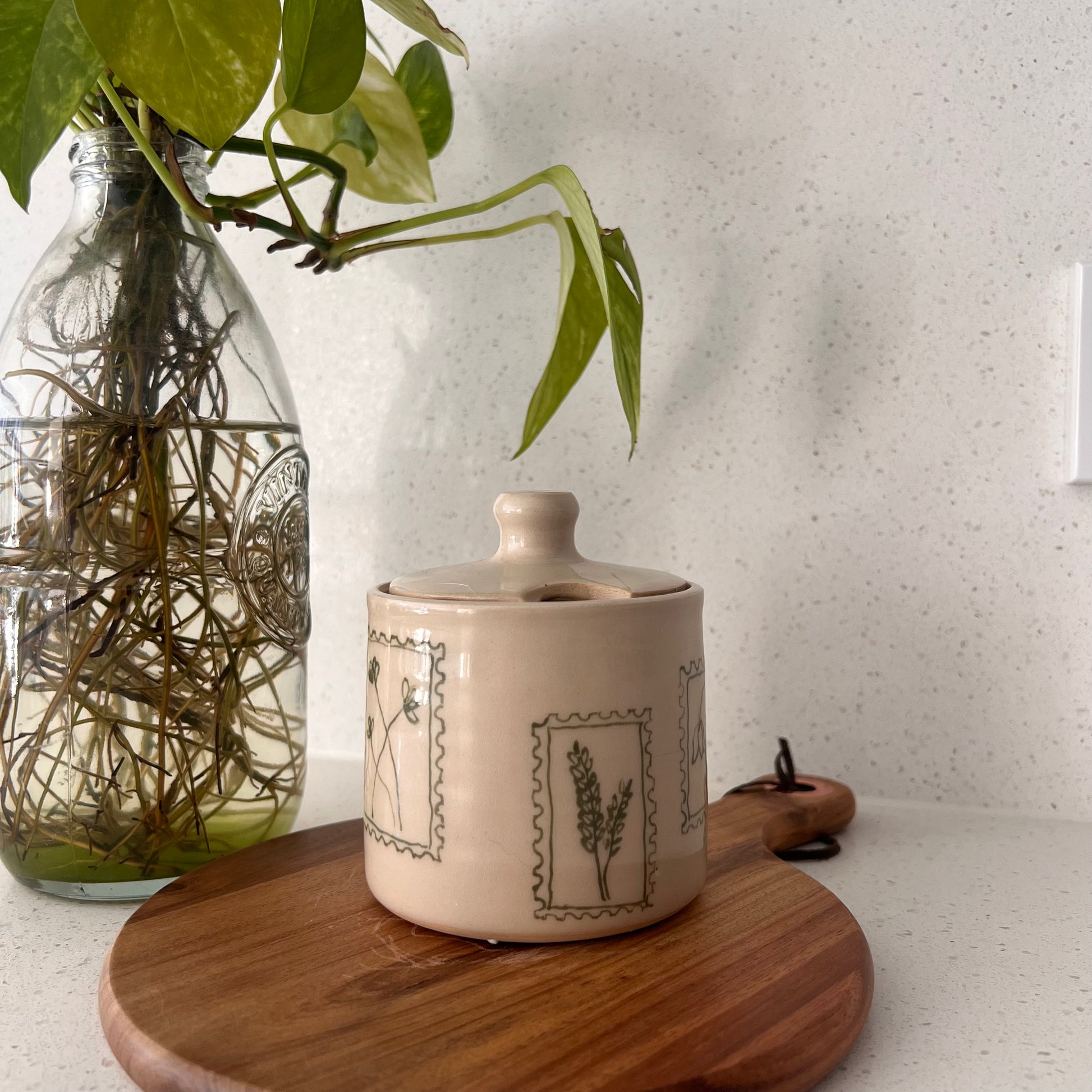Ceramic container with plant designs on a wooden board next to a glass jar with plants.
