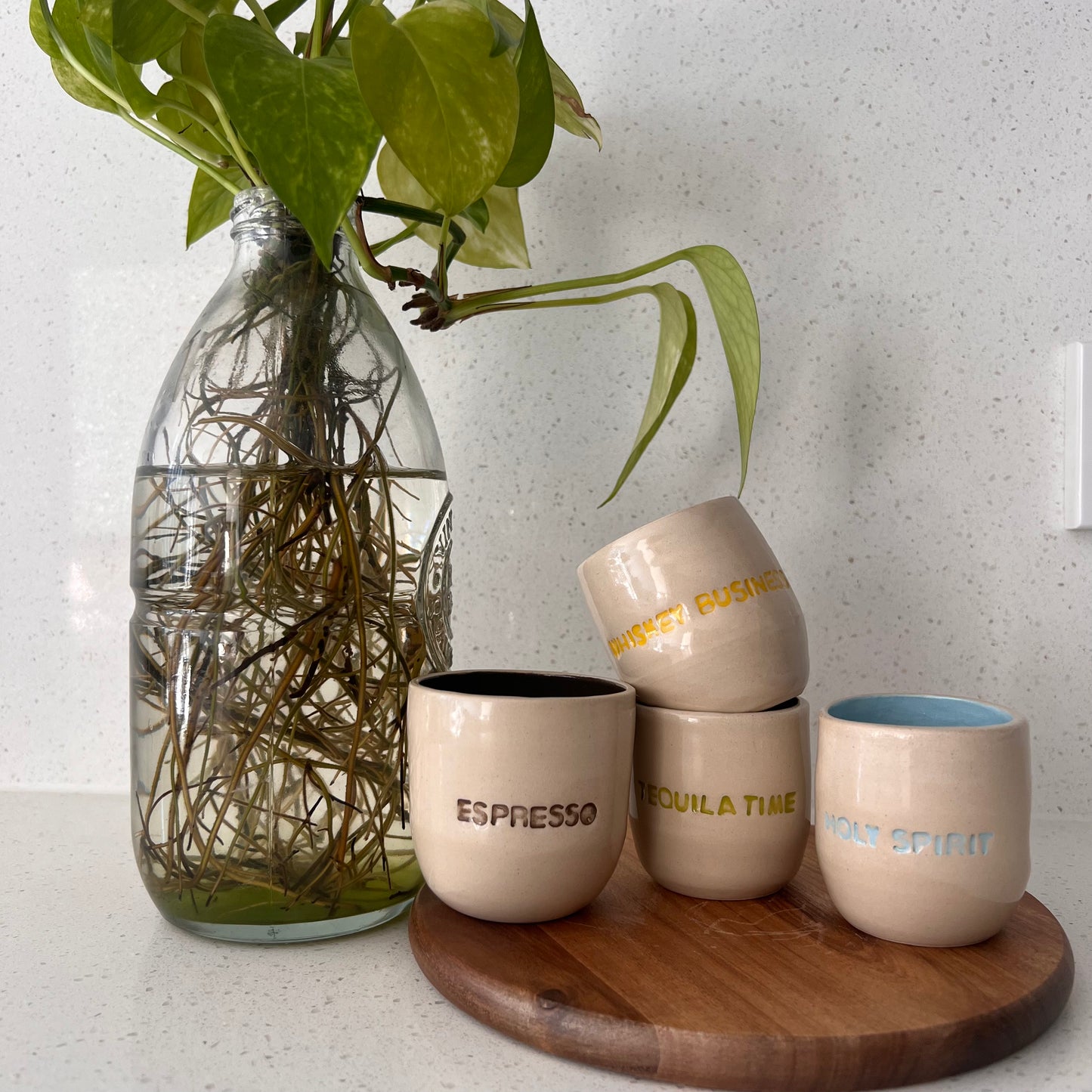 Set of ceramic cups with text on a wooden board next to a glass vase with plants.