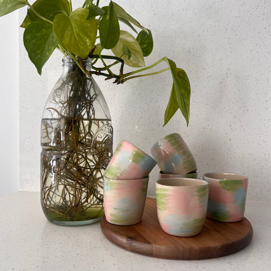 Set of ceramic cups with pastel patterns on a wooden tray, next to a glass vase with green leaves.