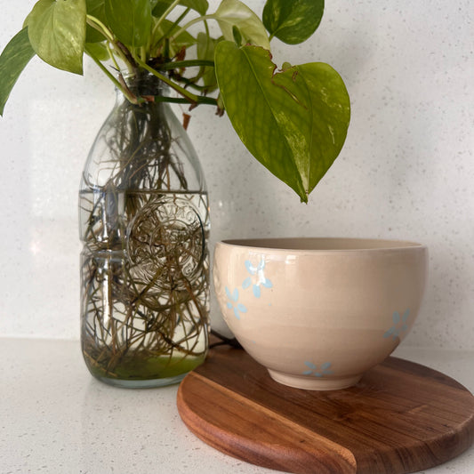 Beige ceramic bowl with blue floral patterns on a wooden coaster, next to a glass vase with green leaves.