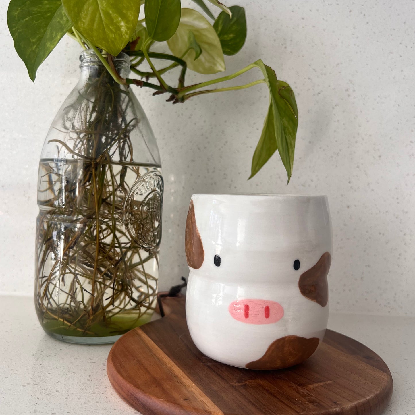 White mug with brown cow design on a wooden coaster next to a glass vase with greenery.